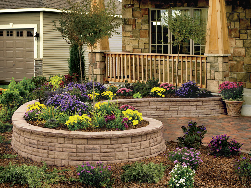 Curved raised Natural Impressions Ashlarstone concrete block planter with purple and yellow flowers in front of a wood and stone porch. 