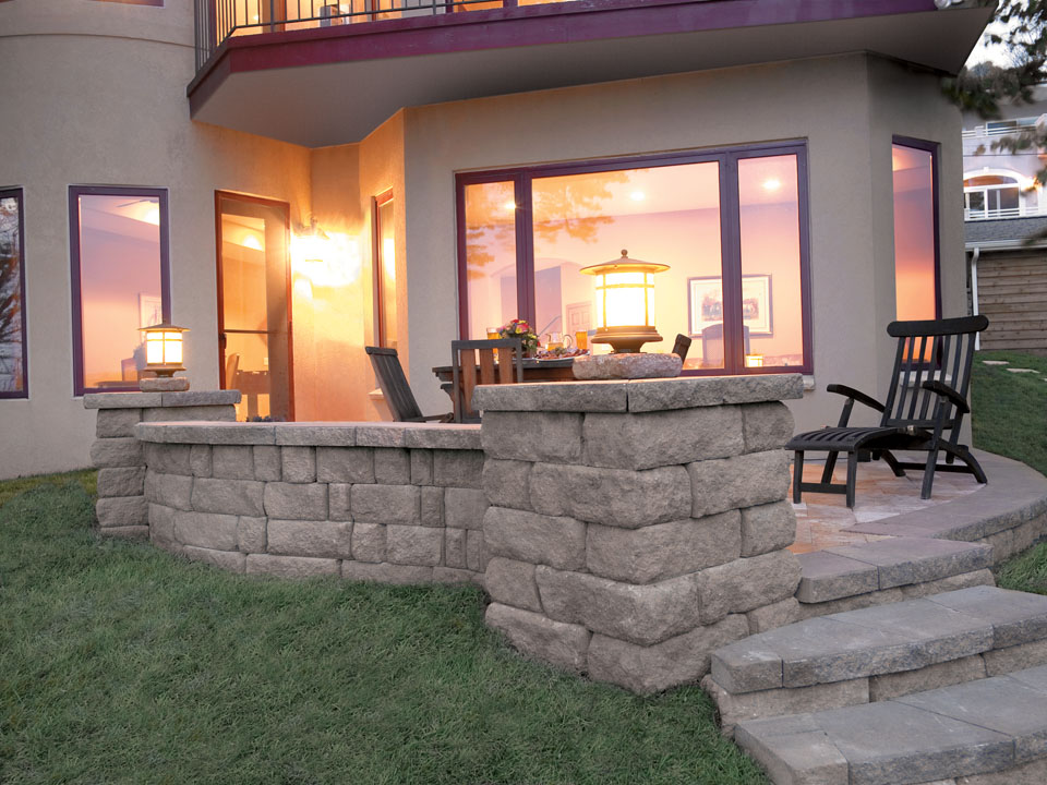 Residential backyard stone paver patio with concrete block walls, lantern-topped capped columns, and stairs